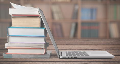 Stapel Bücher auf einem Tisch, daran lehnt ein aufgeklappter Laptop