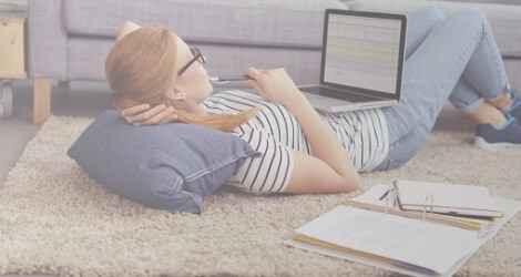 Studentin liegt auf dem Rücken auf dem Boden mit ihrem Laptop auf dem Bauch und Studienmaterial neben sich