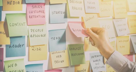 Ein Mann klebt einen Klebezettel an eine Wand zu vielen anderen bunten Klebezetteln, auf denen Schlagwörter aus dem Medien- und Marketingbereich stehen